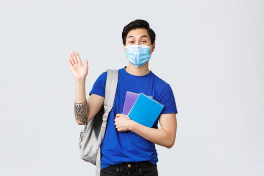 Back to school, studying during covid-19, education and university life concept. Young handsome asian male student, freshman in college waving hand in hello or greeting, holding notebooks.