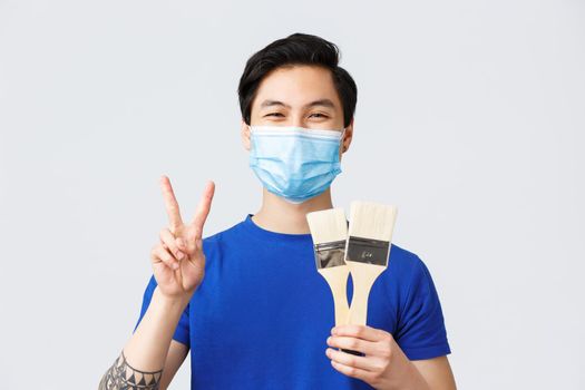 Different emotions, lifestyle and leisure during coronavirus, covid-19 concept. Happy asian man found hobby on self-quarantine showing two brushes, painting and make peace sign, wear medical mask.