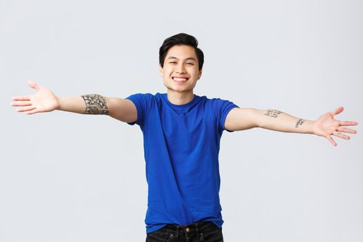 Handsome friendly, smiling asian man, male student in blue t-shirt, spread hands sideways welcoming, greeting friends, want to cuddle or embrace something, standing grey background.