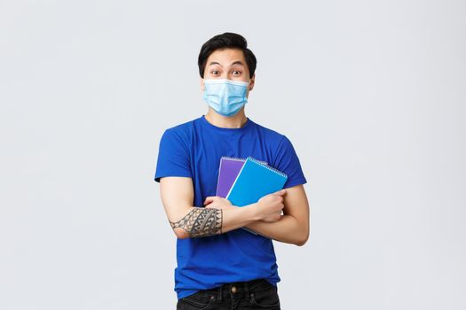 Back to school, studying during covid-19, education and university life concept. Excited happy young male asian student in medical mask heading campus, smiling and holding notebooks.