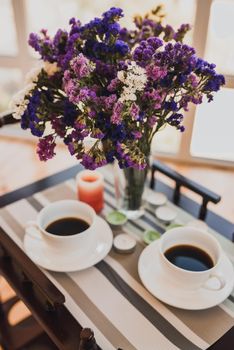 Two cups of coffee sit on a small glass table next to a bouquet of vibrant purple pink flowers in a vase. Breakfast at the hotel.