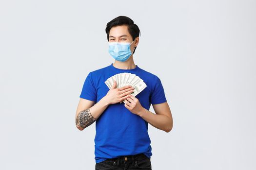 Money, lifestyle, insurance and investment concept. Satisfied griedy asian guy hugging cash of dollars and smiling pleased, wearing medical mask, earned or won lottery.