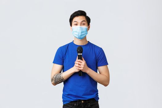 Covid019 lifestyle, people emotions and leisure on quarantine concept. Cute young asian man in medical mask want sing song, holding microphone and looking upbeat camera, singing karaoke.