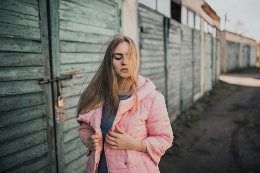 Young gorgeous blonde girl dressed fashion pink jacket and blue jeans. Old azure cerulean fence on the background. Picture ideal for illustrating women's magazines.