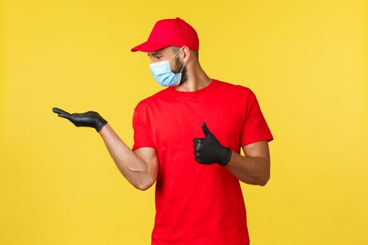 Express delivery during pandemic, covid-19, safe shipping, online shopping concept. Pleased courier in red uniform cap, t-shirt and medical mask show thumb-up and looking at palm satisfied.