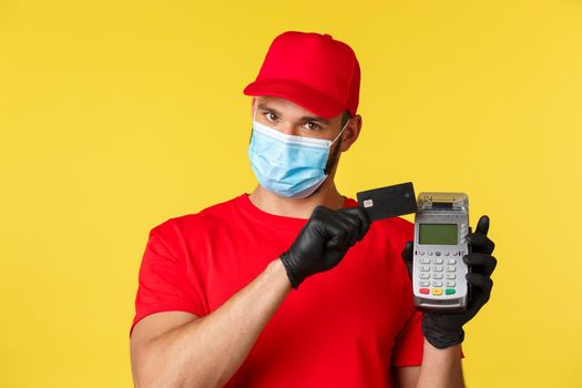 Covid-19, delivery orders, shopping, contactless payment and social distancing concept. Close-up of handsome courier in red uniform and medical mask, press credit card to POS terminal.
