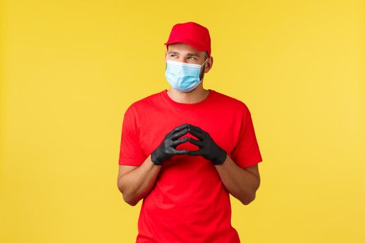 Express delivery during pandemic, covid-19, safe shipping, online shopping concept. Sly and thoughtful courier in red uniform, gloves and medical mask, thinking, steeple fingers, look away.