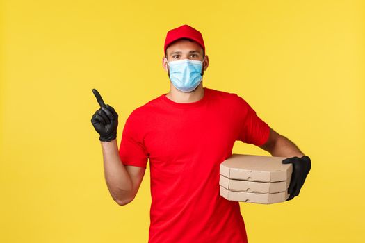 Food delivery, tracking orders, covid-19 and self-quarantine concept. Courier in red uniform and medical mask, provide contactless fast deliver, pointing finger left, holding pizza.