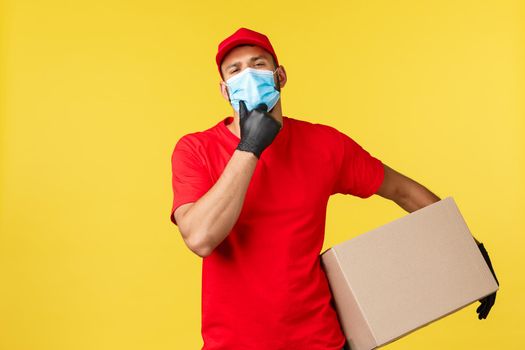 Express delivery during pandemic, covid-19, safe shipping, online shopping concept. Handsome courier in red uniform and medical mask, holding package, touch chin sassy, yellow background.