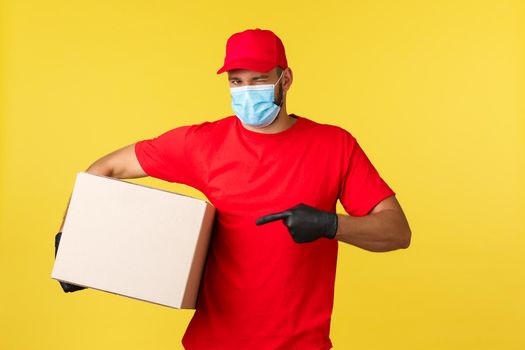 Express delivery during pandemic, covid-19, safe shipping, shopping concept. Cheeky handsome courier bring your parcel, wink and pointing box, wear medical mask protecting from coronavirus spread.