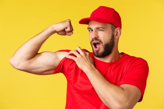 Cheerful handsome delivery guy in red uniform cap and t-shirt, winking at camera as show-off with strong muscles, flex biceps ensure can carry any parcel, courier brag strength over yellow background.