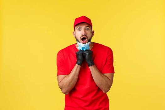Express delivery during pandemic, covid-19, safe shipping, online shopping concept. Shocked and startled courier in red uniform, gloves, take-off medical mask and open mouth saying wow.