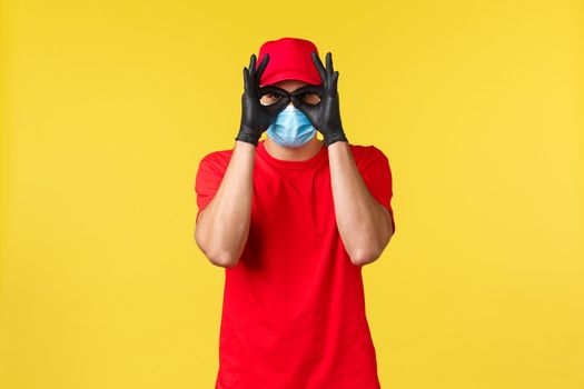 Express delivery during pandemic, covid-19, safe shipping, online shopping concept. Funny courier in red uniform, medical mask and gloves, show okays or glasses with hands.
