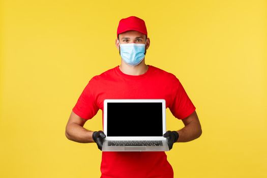 Express delivery during pandemic, covid-19, safe shipping, online shopping concept. Employee in red uniform cap and gloves, courier in medical mask showing website on laptop screen.