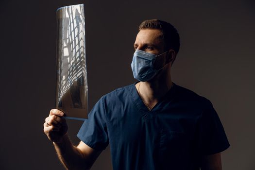 Doctor in medical mask for protection of coronavirus covid-19 holding x-ray scan of ill patient. Computered tomography. Surgeon in medical robe looking at picture of scan on dark background.
