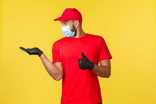 Express delivery during pandemic, covid-19, safe shipping, online shopping concept. Impressed courier, employee in red uniform and medical mask, thumb-up as looking at his hand.