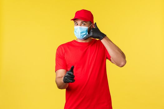 Express delivery during pandemic, covid-19, safe shipping, online shopping concept. Handsome courier in red uniform, medical mask and gloves, pointing client and make phone call sign.