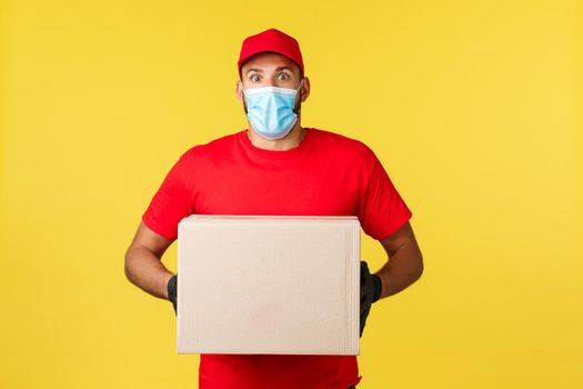 Express delivery during pandemic, covid-19, safe shipping, online shopping concept. Startled and shocked courier in red uniform, medical mask and gloves, holding parcel feel astonished.