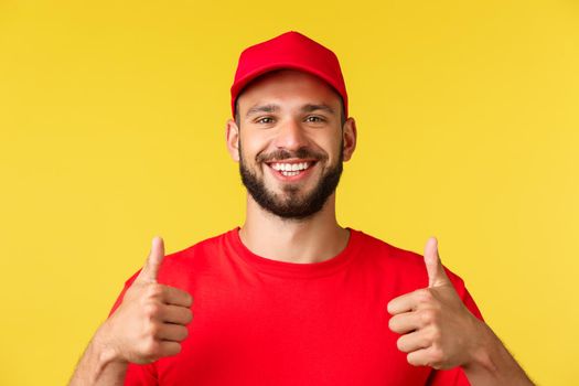 Close-up of friendly smiling delivery guy in red uniform cap and t-shirt provide best express shipping concept, show thumbs-up in approval, recommend using company service, yellow background.