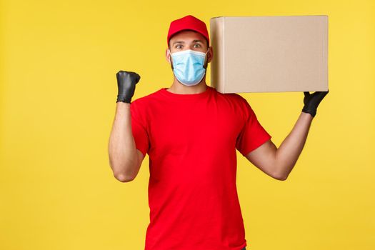 Express delivery during pandemic, covid-19, safe shipping, shopping concept. Rejoicing happy courier fist pump, holding box on shoulder, deliver successfully, chanting saying yes, wear medical mask.