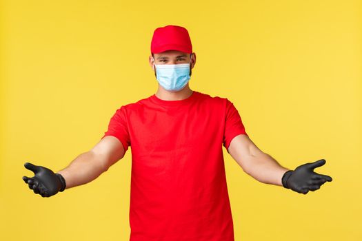 Express delivery during pandemic, covid-19, safe shipping, online shopping concept. Handsome courier in red uniform, medical mask and gloves, spread hands to hold parcel, carry package to clients.