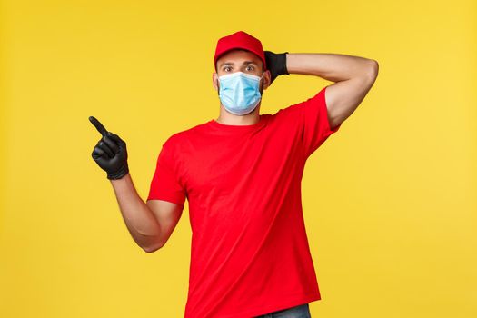 Express delivery during pandemic, covid-19, safe shipping, online shopping concept. Impressed courier in red logistic company uniform, stare and point left excited, yellow background.