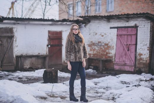 Girl blonde in cat eyes glasses in an artificial faux fur coat posing. Lying snow on the background. Red orange pink tones in the photo. Around the old shabby abandoned buildings. Cold winter