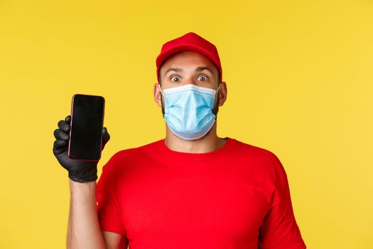Covid-19, delivery orders, shopping, contactless payment and social distancing concept. Excited courier in red uniform, medical mask and gloves, showing excellent promo on mobile phone screen.