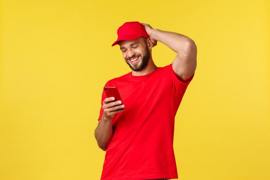 Contactless payment, online delivery, internet shopping and parcel tracking concept. Cheerful courier in red uniform cap and t-shirt, messaging, smiling at smartphone over yellow background.