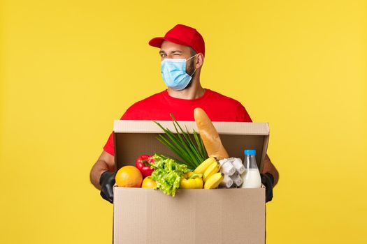 Takeaway delivery, covid-19 quarantine and groceries concept. Customer order online grocery, courier wink at camera bring box full of food, wearing medical mask and red uniform.