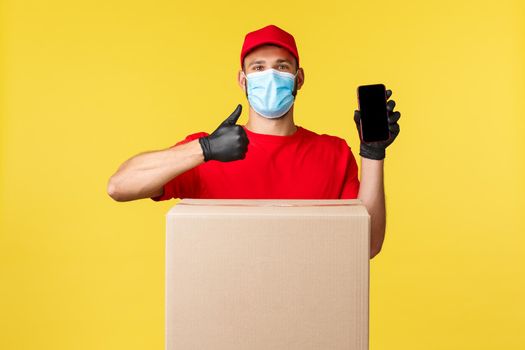 Express delivery during pandemic, covid-19, safe shipping, shopping concept. Courier in red uniform, showing deliver service app, thumb-up, stand near box package, application for tracking order.