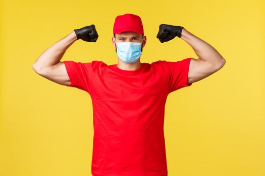 Express delivery during pandemic, covid-19, safe shipping, online shopping concept. Determined young courier in red uniform, medical mask, show-off strength, fix biceps, provide quality service.
