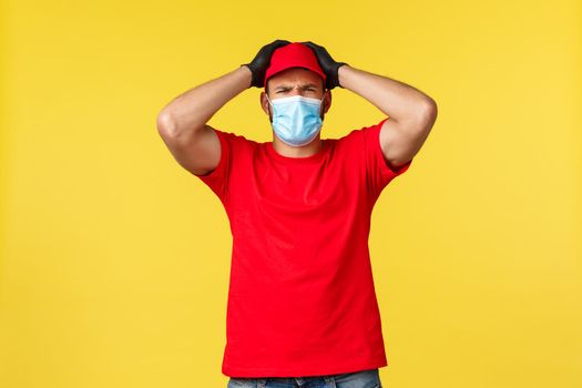 Express delivery during pandemic, covid-19, safe shipping, online shopping concept. Troubled and nervous courier in red uniform, medical mask, grab head have problem, yellow background.