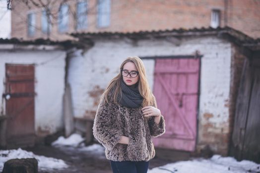 Girl blonde in cat eyes glasses in an artificial faux fur coat posing. Lying snow on the background. Red orange pink tones in the photo. Around the old shabby abandoned buildings. Cold winter
