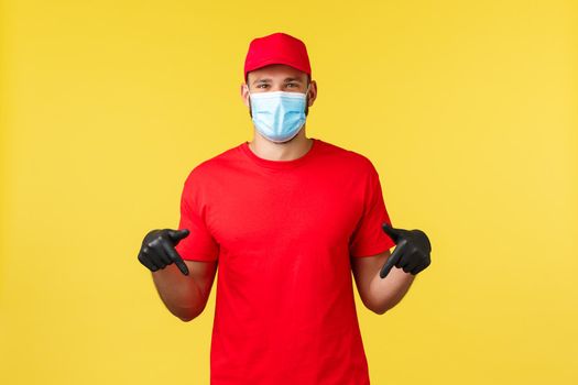 Express delivery during pandemic, covid-19, safe shipping, online shopping concept. Smiling friendly courier, delivery guy in uniform red cap and t-shirt, medical mask, pointing down.