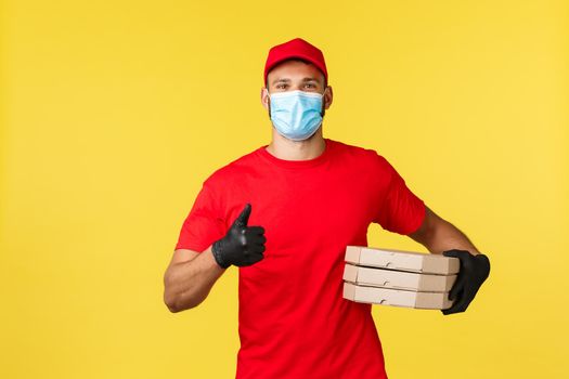 Express delivery during pandemic, covid-19, safe shipping, shopping concept. Friendly young courier in red uniform and medical mask, provide safe order delivery, holding pizzas and show thumb-up.