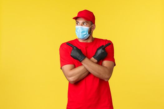 Express delivery during pandemic, covid-19, safe shipping, online shopping concept. Excited and surprised courier in red uniform, medical mask and gloves, look left interested, point sideways.