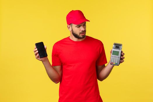 Contactless payment, online delivery, internet shopping concept. Confused young courier in red uniform, newbie courier figure out how POS terminal works, holding mobile phone.