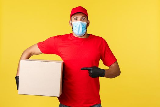 Express delivery during pandemic, covid-19, safe shipping, shopping concept. Shocked gasping courier staring camera speechless as pointing at box, carry package, wrong adress, yellow background.