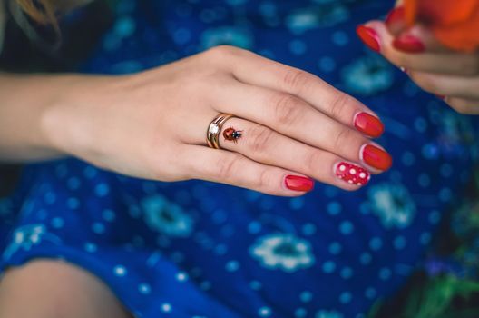 Hand of a girl with fair skin with two rings on a ring finger with red polka dots manicure.On the hand a ladybug.the other hand the girl holds a red poppy flower. Women's blue dress with a daisy print