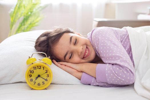 A adorable smiling brunette girl in purple polka dot pajamas lie on a white pillow, on a bed, in the morning light, a yellow alarm clock stands nearby. copy space