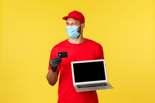 covid-19, safe shopping, express delivery and order tracking concept. Cheerful young male courier in red uniform and medical mask, recommend use contactless payment, show credit card and laptop.