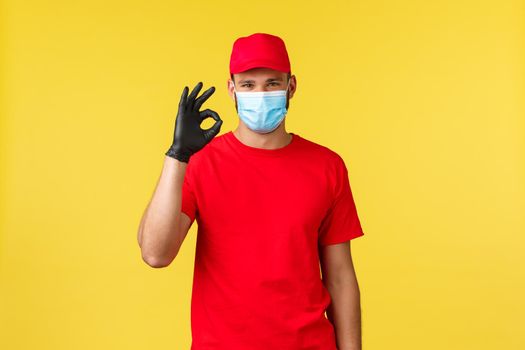 Express delivery during pandemic, covid-19, safe shipping, online shopping concept. Satisfied handsome courier, red uniform and medical mask, provide fast transfer of client orders, show okay sign.