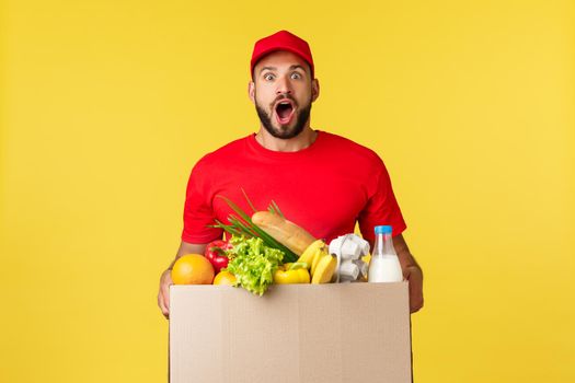 Online delivery, shopping and food order concept. Excited and amused courier in red uniform, bearde delivery man gasping astonished, bring grocery box to customer home, yellow background.