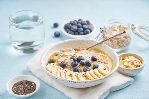 Oatmeal with blueberries, banana and cup of water on blue light background. Side view. Healthy diet breakfast