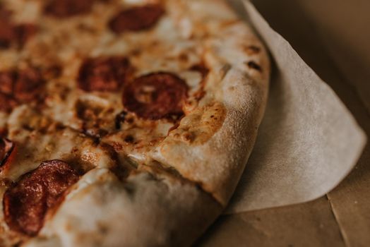 A large round juicy mouth-watering pizza with cheese and salami lies on a cardboard box from a delivery package. paper backing. close view. see flour. good thin dough