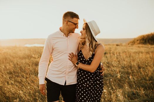 a guy with a girl in hat walking in the meadow. A couple of fair-haired fair-skinned people in love are resting in nature in a field at sunset.