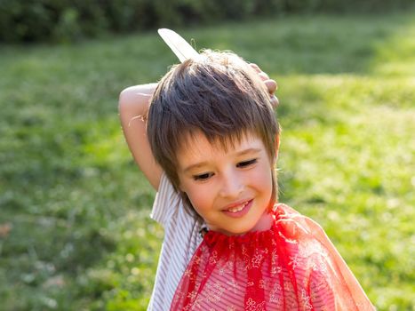 Portrait of smiling boy playing American Indian. Kid with white bird feather and red cloak. Costume role play. Outdoor leisure activity.