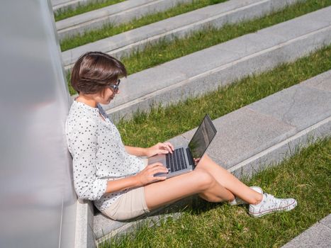 Woman sits with laptop on urban park bench. Freelancer at work. Student learns remotely from outdoors. Modern lifestyle. Summer vibes. Outdoor workplace.