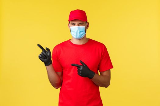 Express delivery during pandemic, covid-19, safe shipping, online shopping concept. Young friendly courier in red uniform, medical mask and gloves, pointing fingers left, wink at client.